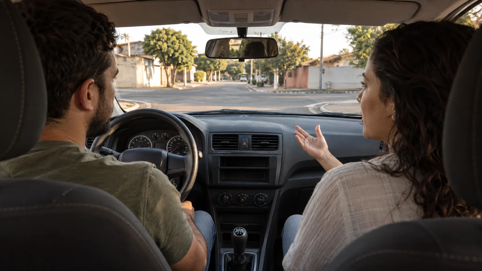 Duas pessoas conversam dentro de um carro parado enquanto observam um cruzamento à frente.