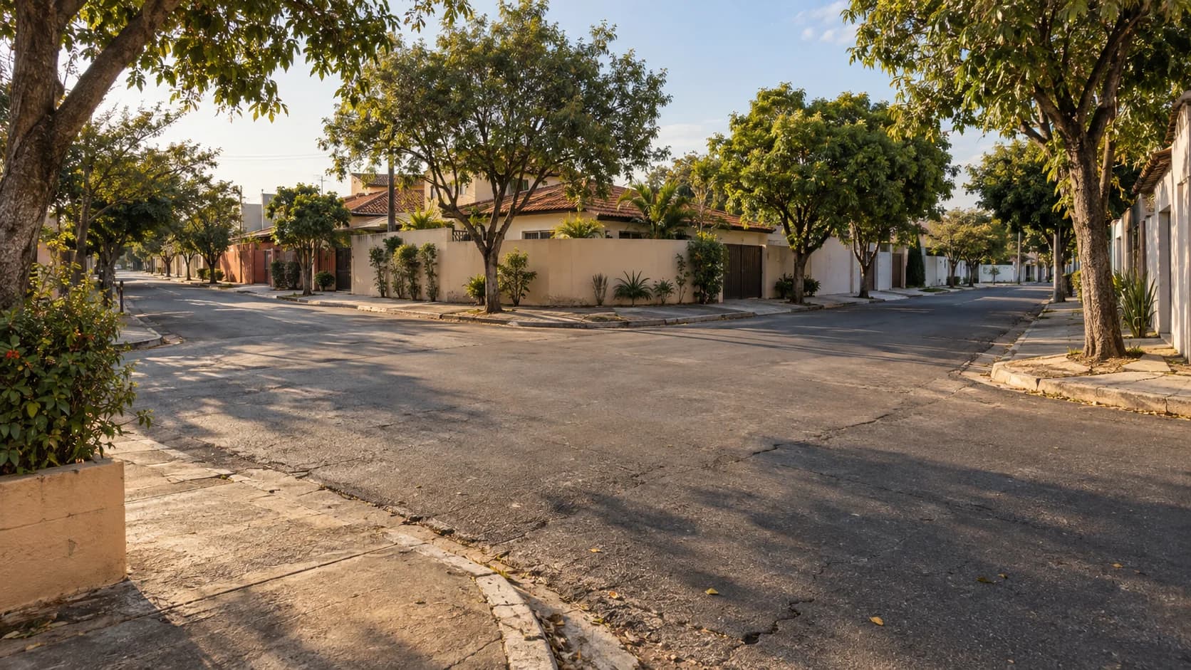Cruzamento residencial vazio, com ruas sem sinalização e casas arborizadas sob a luz do fim da tarde.