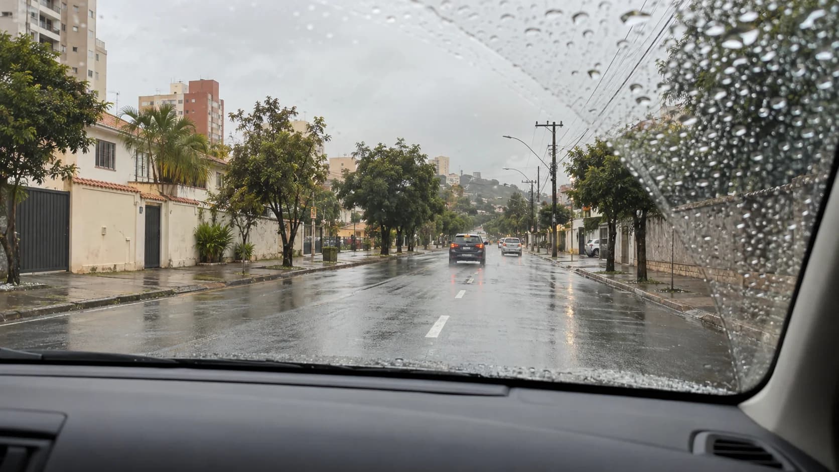 Rua urbana molhada vista pelo para-brisa com gotas de chuva e amplo espaço até os veículos à frente.