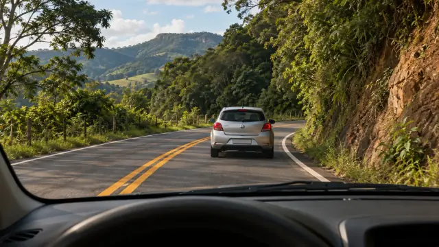 Pelo para-brisa, um carro segue à frente numa estrada de duplo sentido; uma curva contorna uma parede de rocha e a linha central é amarela contínua.